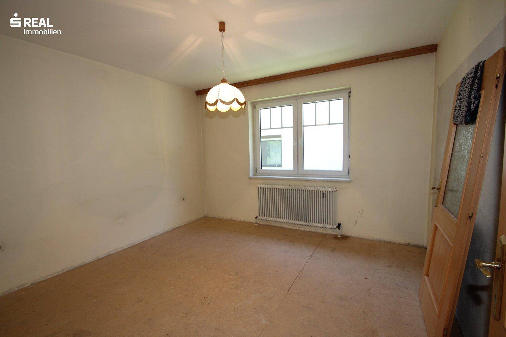 Schlafzimmer mit Teppichboden, großem Fenster und einfacher Deckenleuchte, renovierungsbedürftig.