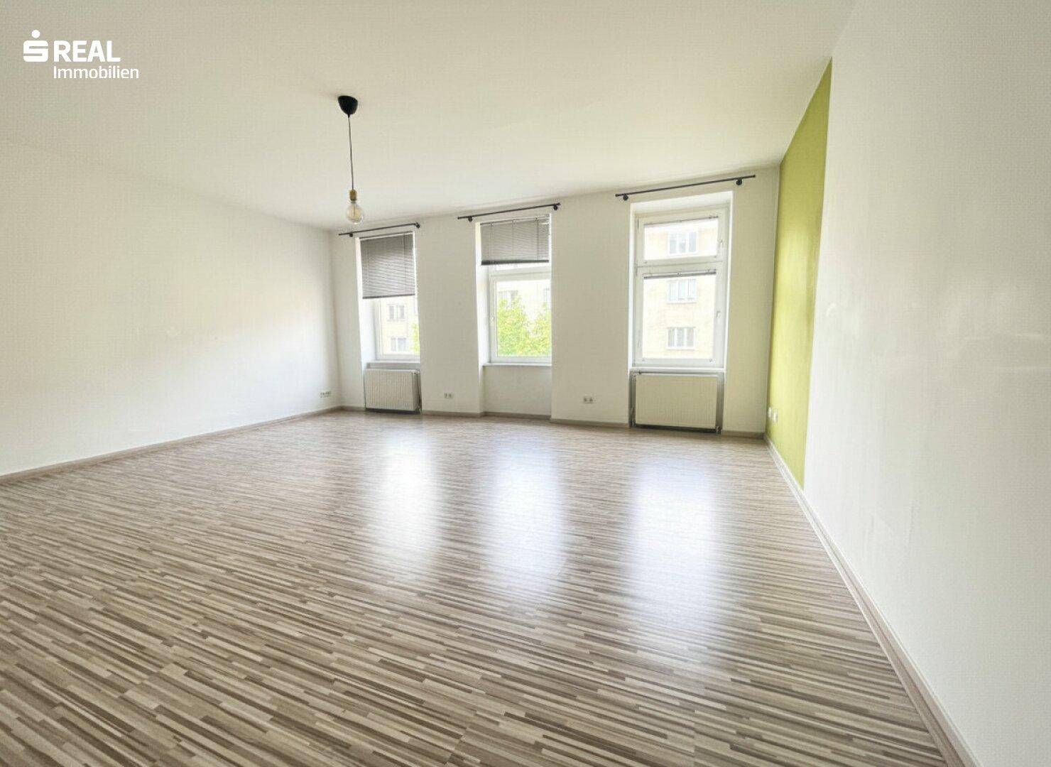 Heller, leerer Wohnraum mit Laminatboden, großen Fenstern und einer Akzentwand in Lindgrün.