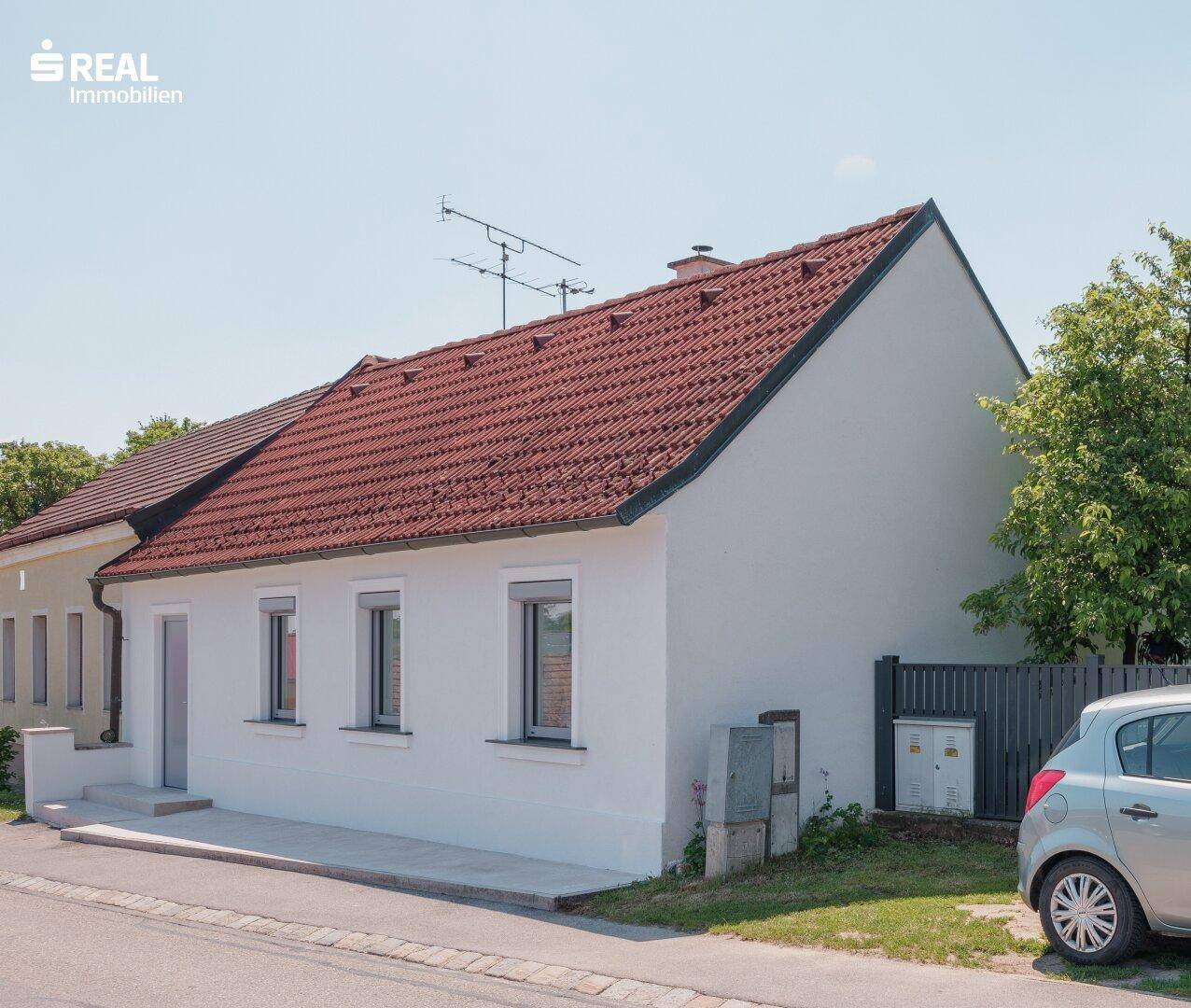 Seitenansicht des Hauses von der Straße mit rotem Ziegeldach und Fenstern an einem sonnigen Tag.