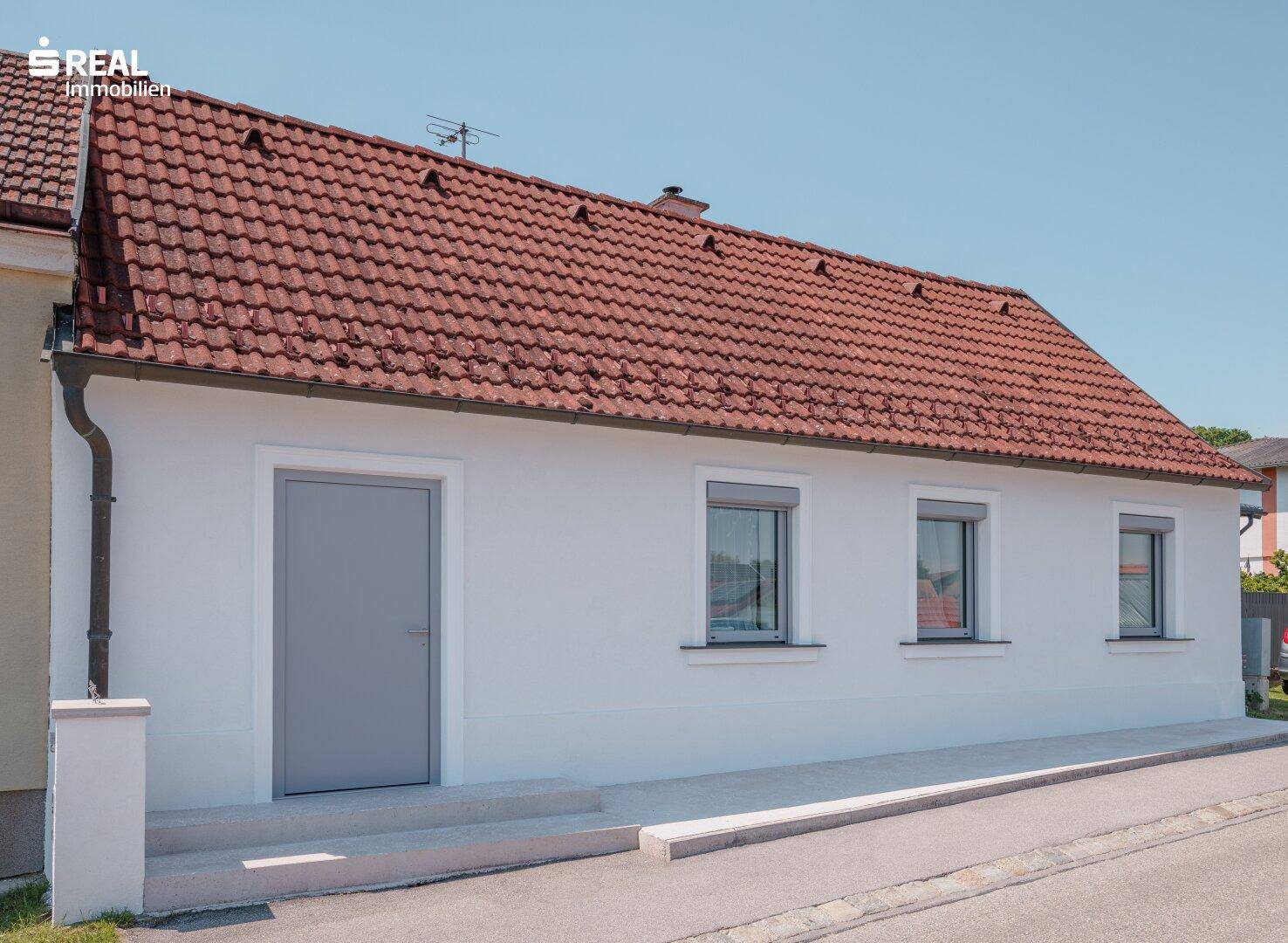 Vorderansicht des Hauses mit grauer Eingangstür, Rollläden und rotem Ziegeldach unter klarem Himmel.