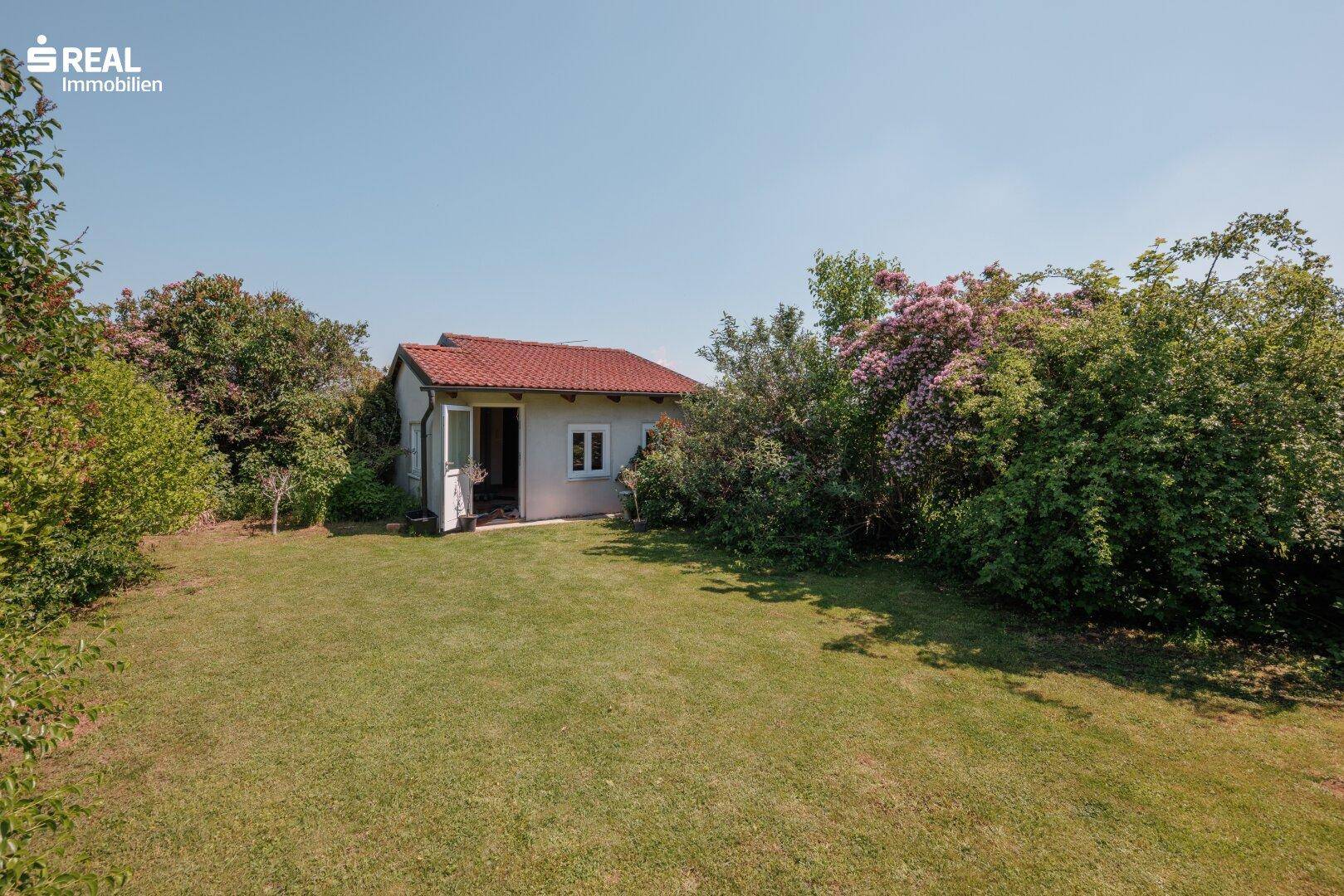 Kleines Gartenhaus umgeben von viel Grün und einer weitläufigen Rasenfläche unter blauem Himmel.