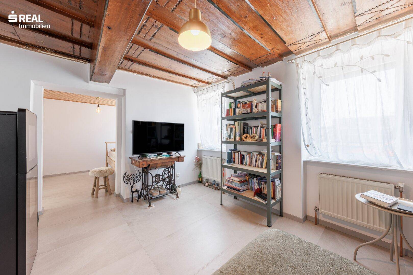 Helles Wohnzimmer mit Holzbalkendecke, großem Fenster und Bücherregal, das zum Verweilen einlädt.