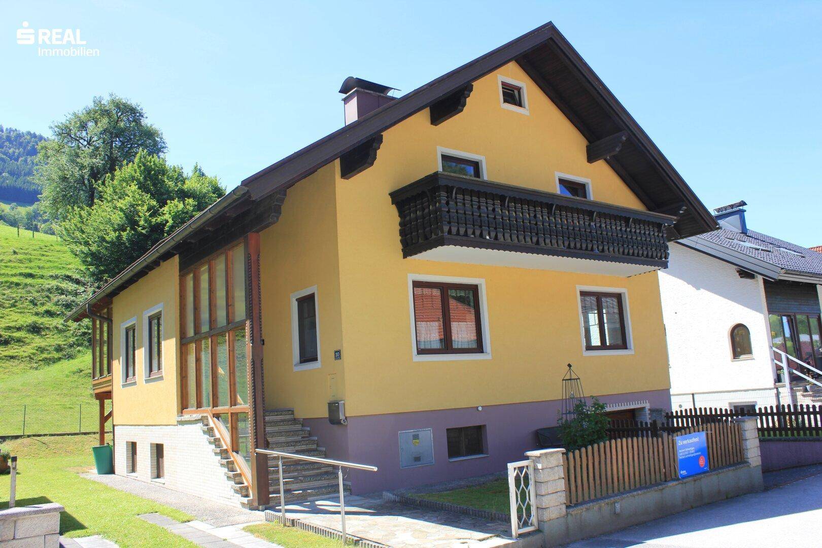 Ansicht des gelben Hauses mit verglastem Anbau und traditionellem Holzbalkon in ländlicher Umgebung.