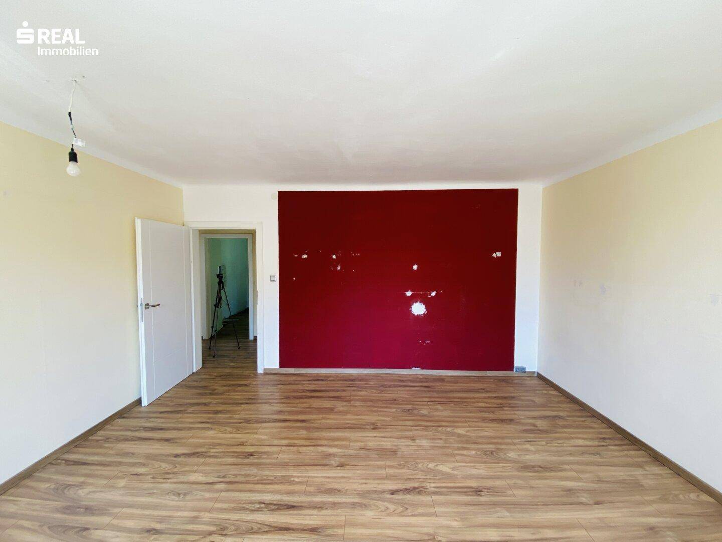 Geräumiges Zimmer mit Laminatboden und einer roten Akzentwand, die Renovierungsbedarf aufweist.