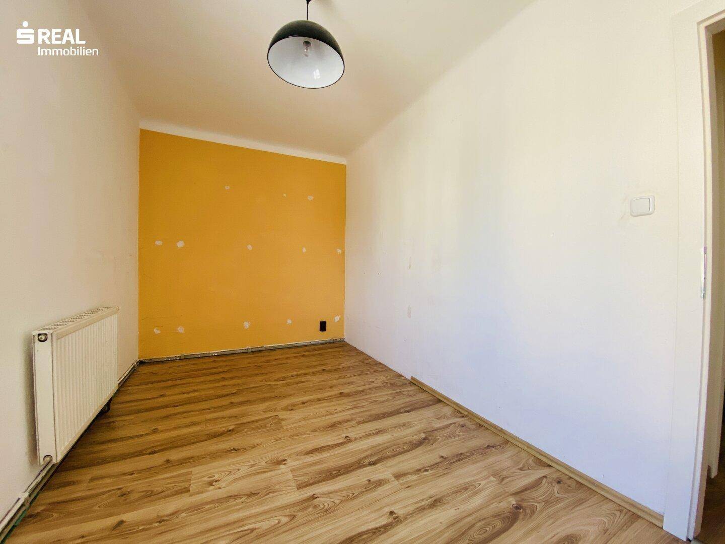 Kleines Zimmer mit Laminatboden, einer gelben Akzentwand und einem Heizkörper.
