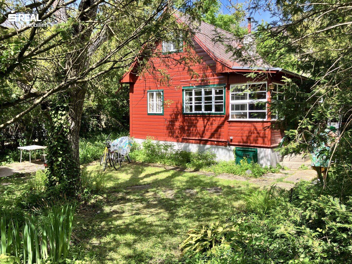 Rotes Holzhaus mit grünen Fensterrahmen, umgeben von üppiger Vegetation und Bäumen.