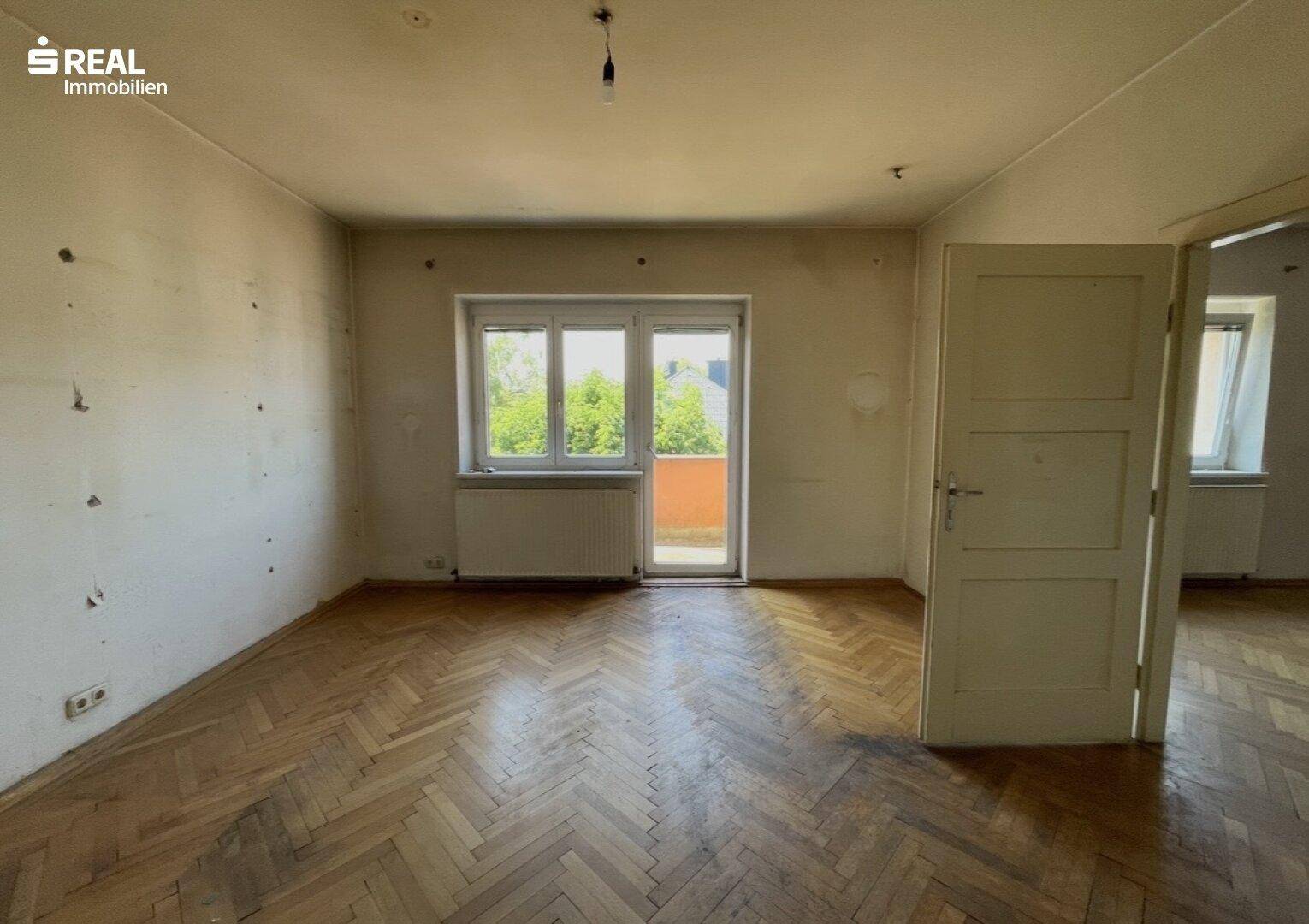 Geräumiges Zimmer mit Fischgrätparkett, Fenster und Zugang zu einem Balkon, renovierungsbedürftig.