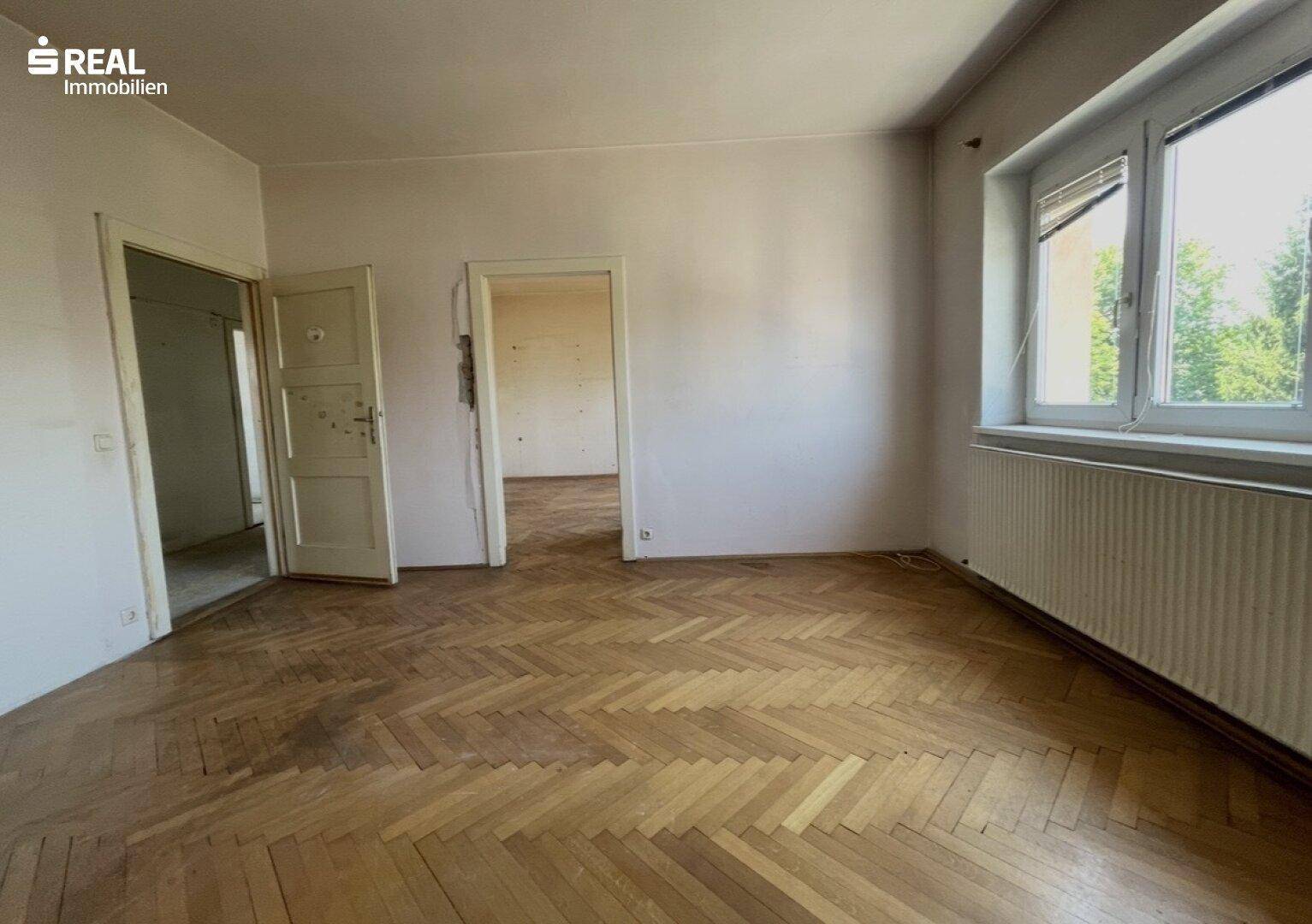 Helles Zimmer mit klassischem Fischgrätparkett, großem Fenster und Zugang zu angrenzenden Räumen.