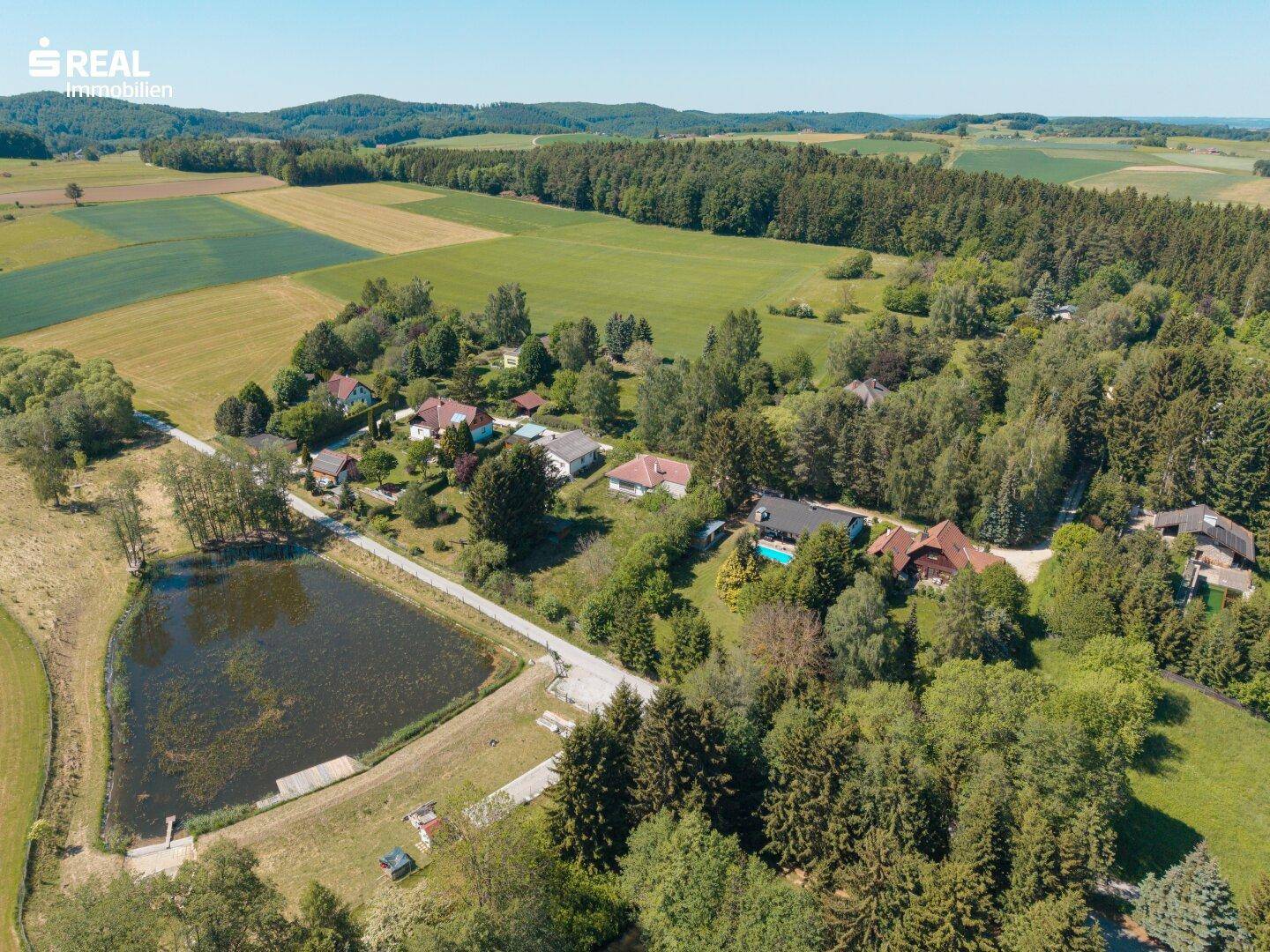 Weitläufige Luftaufnahme der ländlichen Umgebung mit Teich, Feldern, Wäldern und Siedlung.