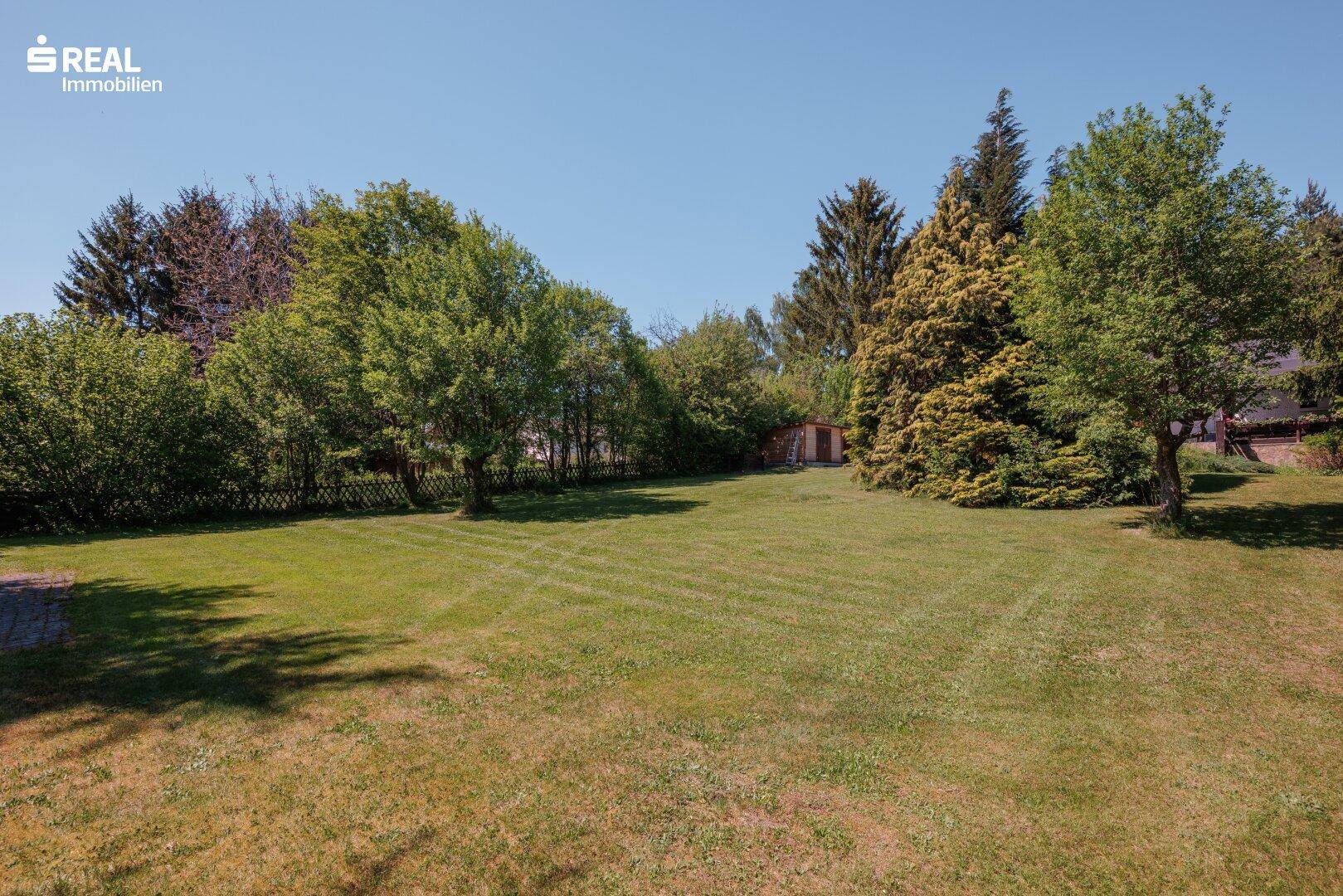 Große, gepflegte Rasenfläche mit Bäumen und einem Gartenhaus in der Ferne.