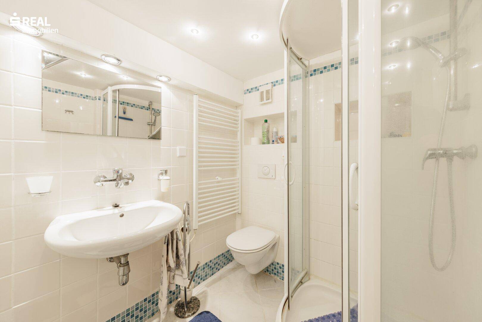 Helles Badezimmer mit Eckdusche, Waschbecken und WC, ausgestattet mit einem Handtuchheizkörper.