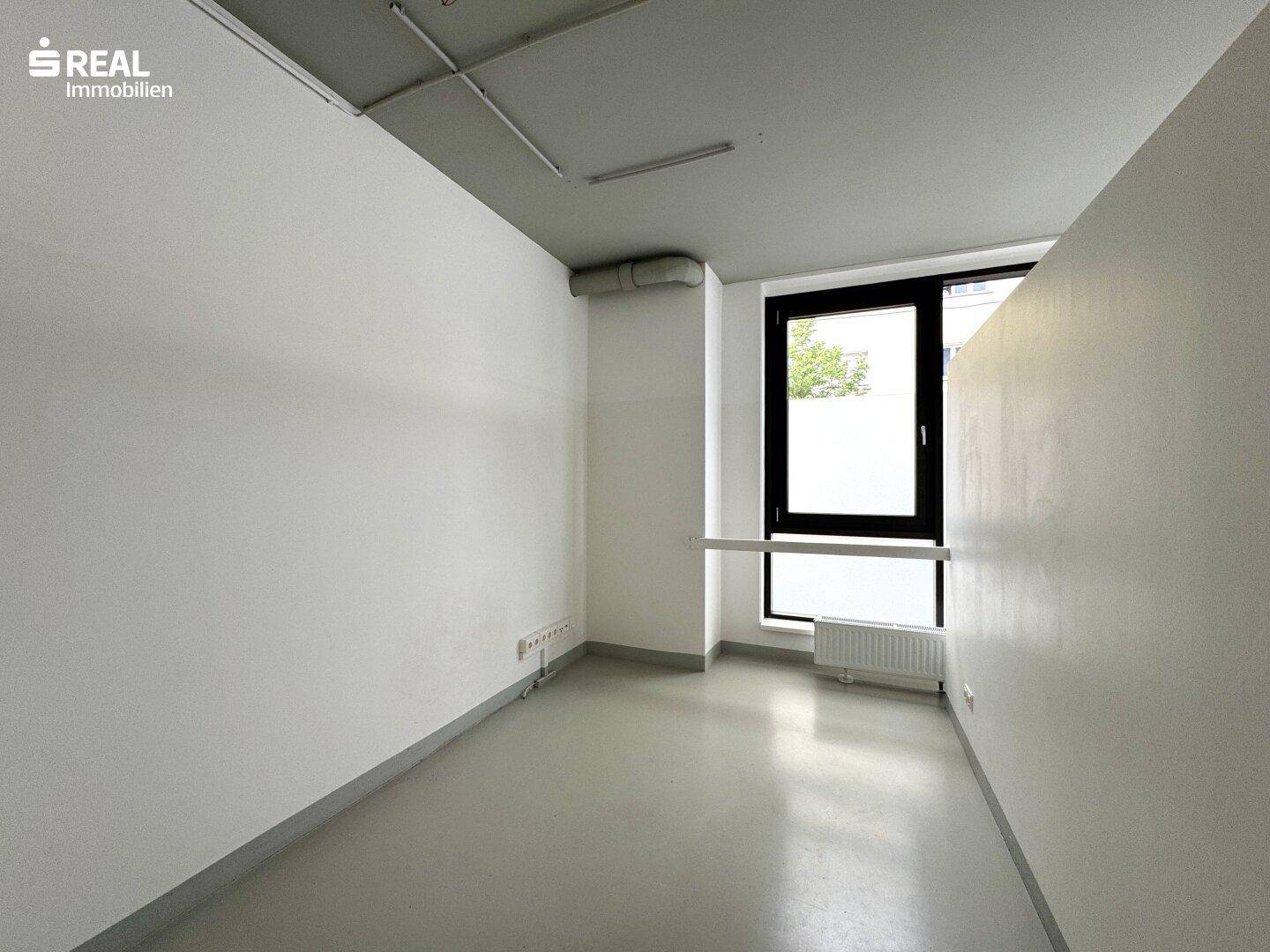 Kleiner Büroraum mit einem Fenster, das natürliches Licht hereinlässt, und einem Heizkörper darunter.