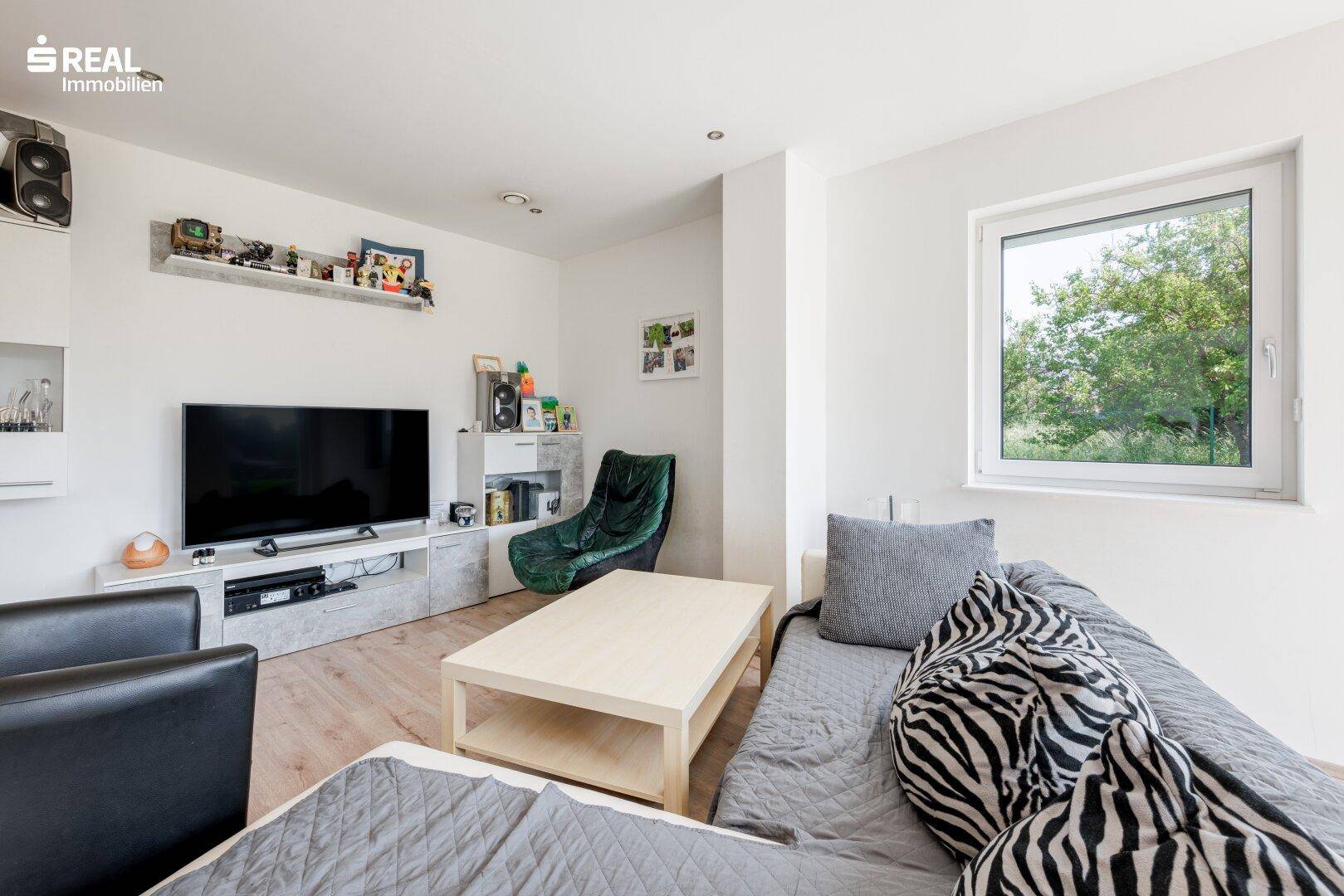 Gemütliches Wohnzimmer mit Sofa, Fernseher und Fenster mit Blick ins Grüne.