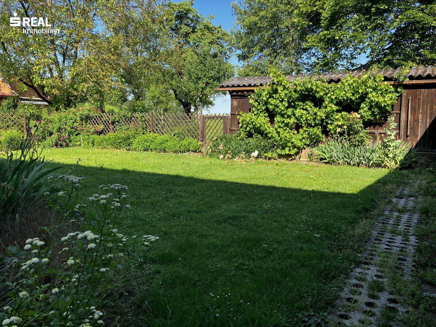 Gepflegter Rasenbereich im Garten mit Bäumen und Sträuchern, umgeben von einem Holzzaun.
