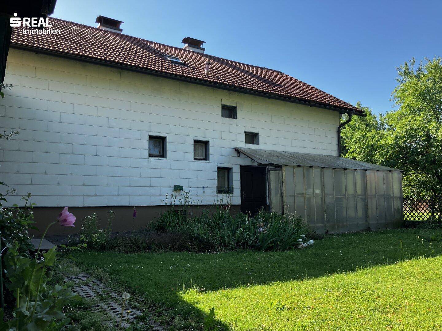 Rückseite des Hauses mit einem kleinen Gewächshaus und Gartenbereich unter blauem Himmel.