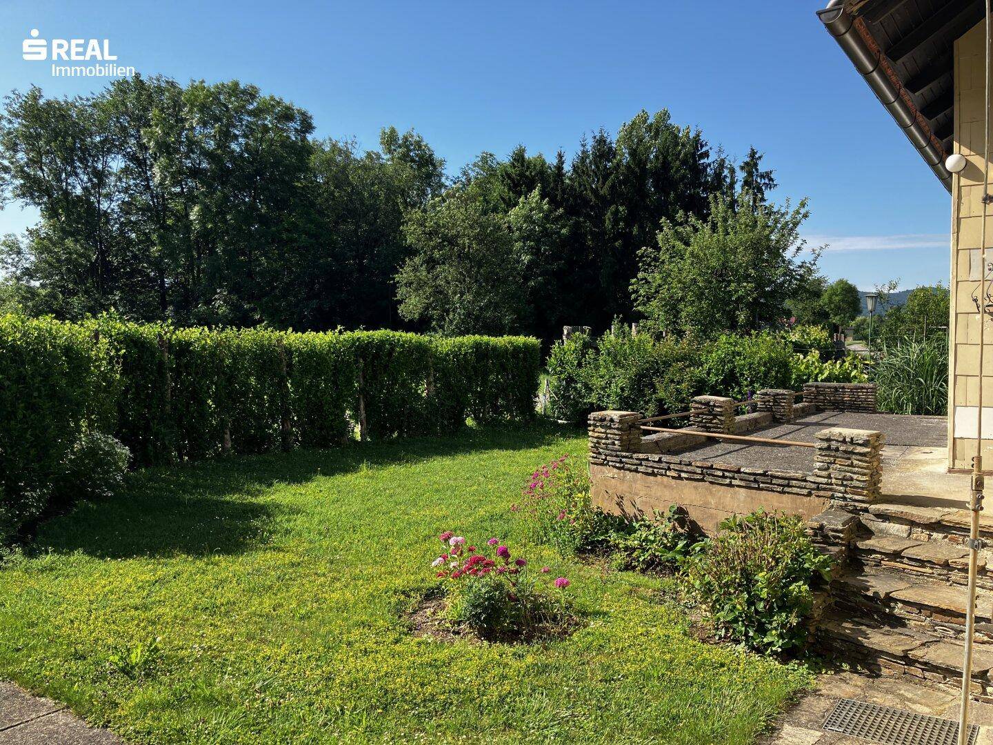 Terrasse mit Steinstufen und angrenzendem Rasenbereich, umgeben von einer hohen Hecke.