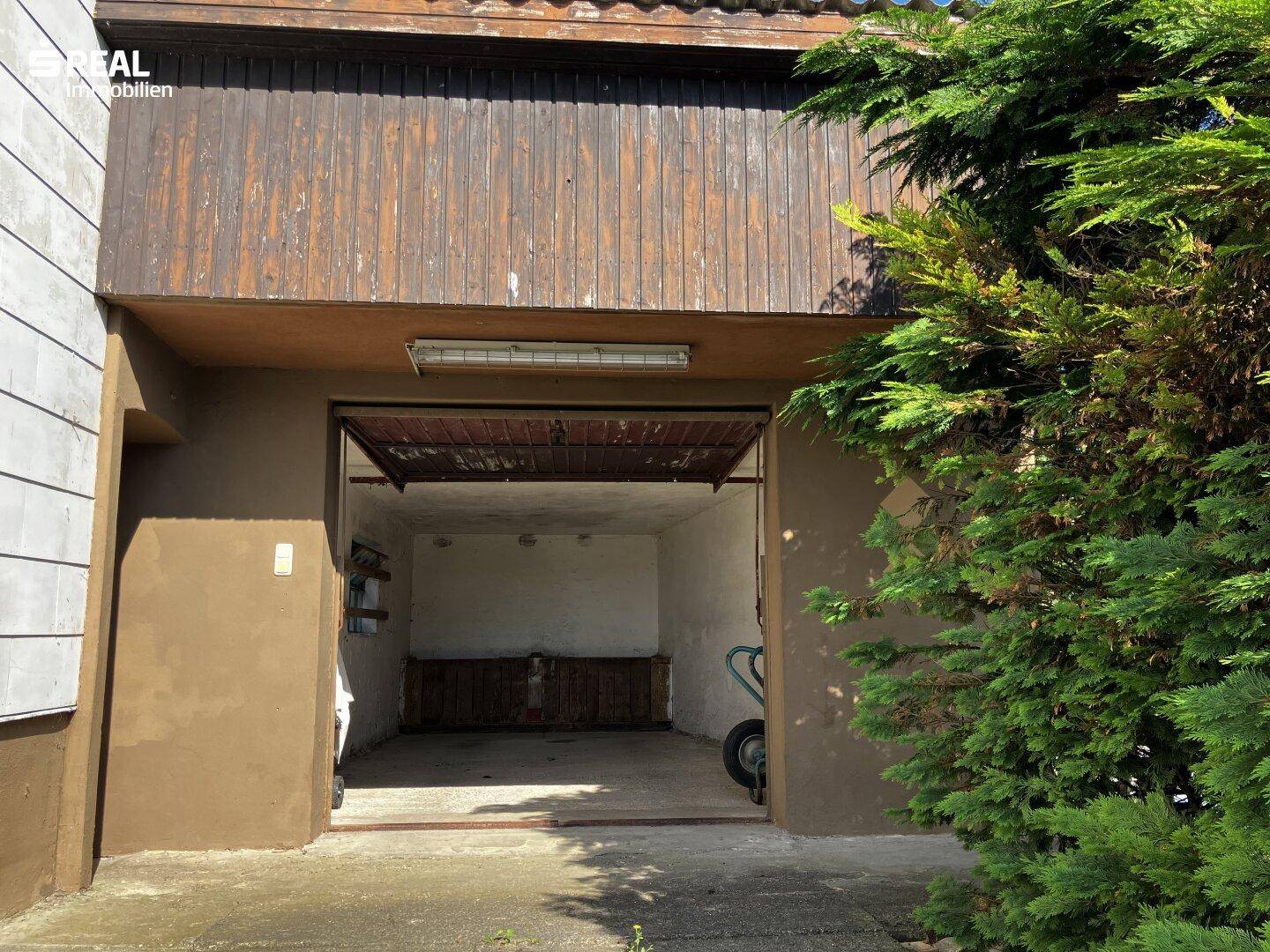 Offene Garage mit Holztor und Werkbank im Inneren, umgeben von Grünpflanzen.