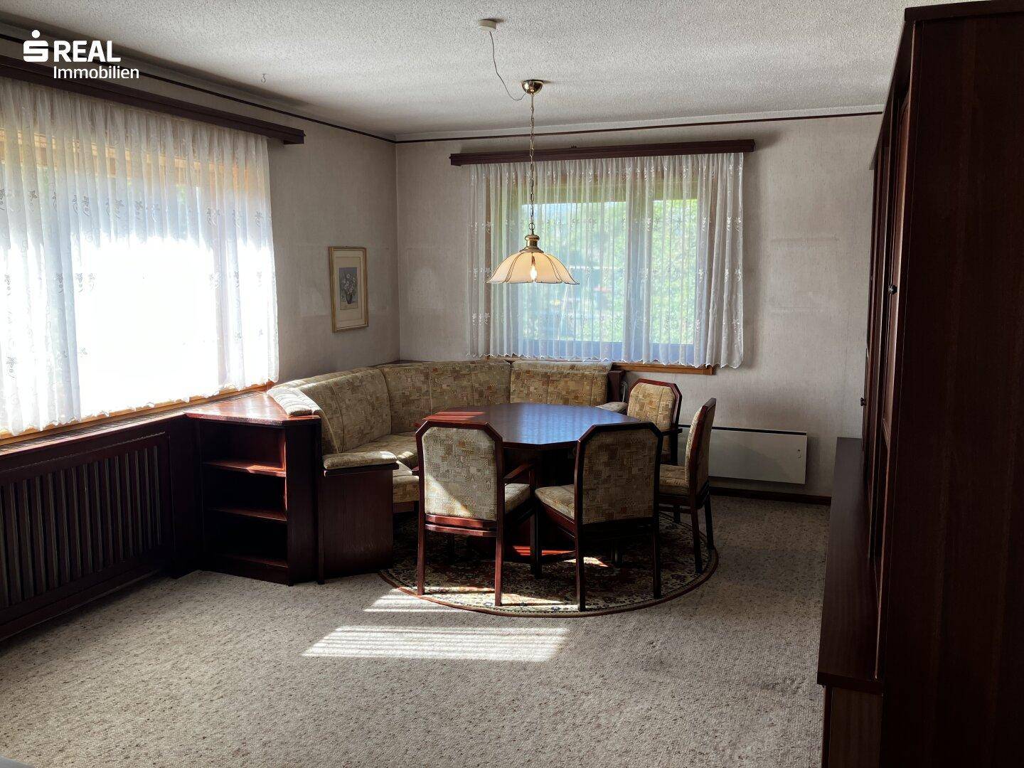 Gemütliches Wohnzimmer mit Teppichboden, Eckbank und Esstisch, umgeben von großen Fenstern.