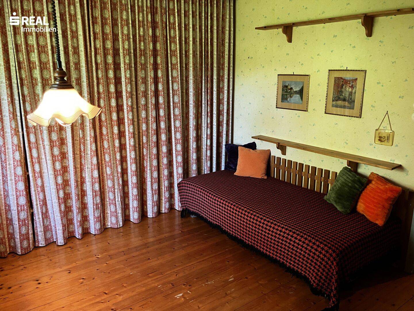 Schlafzimmer mit Holzboden, Tagesbett und gemusterten Vorhängen, hell durch Fenster.
