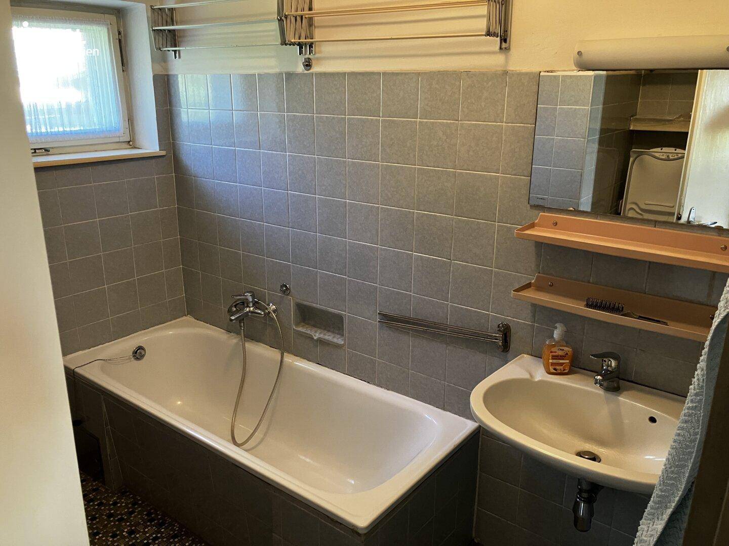 Badezimmer mit grauen Fliesen, Badewanne und Waschbecken unter einem Fenster.