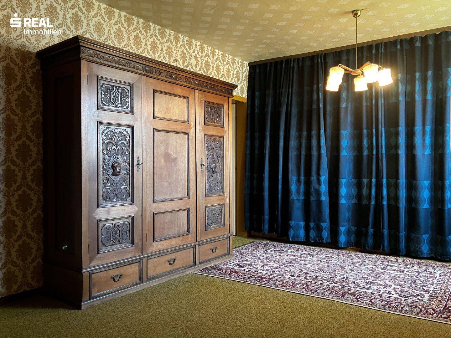 Schlafzimmer mit großem, geschnitztem Holzschrank, Teppichboden und dunklen Vorhängen.