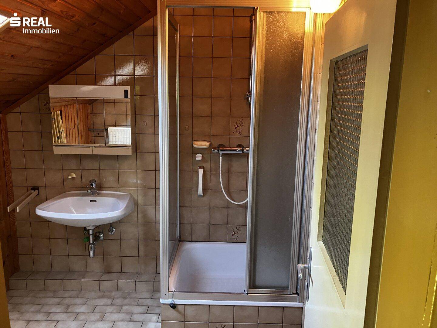 Badezimmer mit braunen Fliesen, Duschkabine und Waschbecken unter Dachschräge.