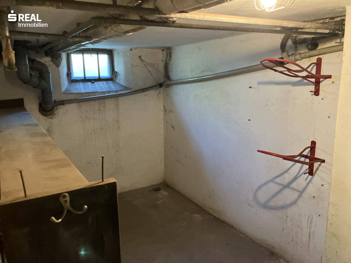 Weiterer Kellerraum mit sichtbaren Rohren und einfachen Wandhaken für Lagerzwecke.