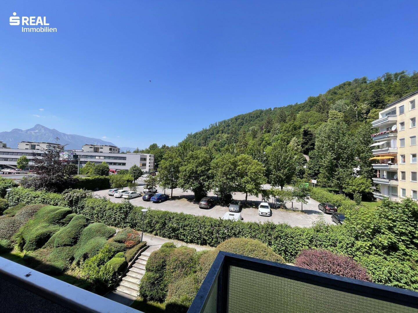 Weitläufiger Blick vom Balkon auf grüne Bäume, Parkplätze und die umliegende Berglandschaft.