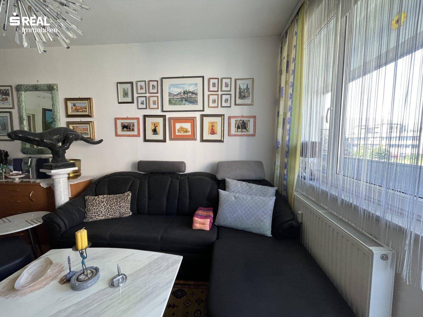 Gemütliches Wohnzimmer mit dunklem Sofa, vielen Bildern an der Wand und großem Fenster.