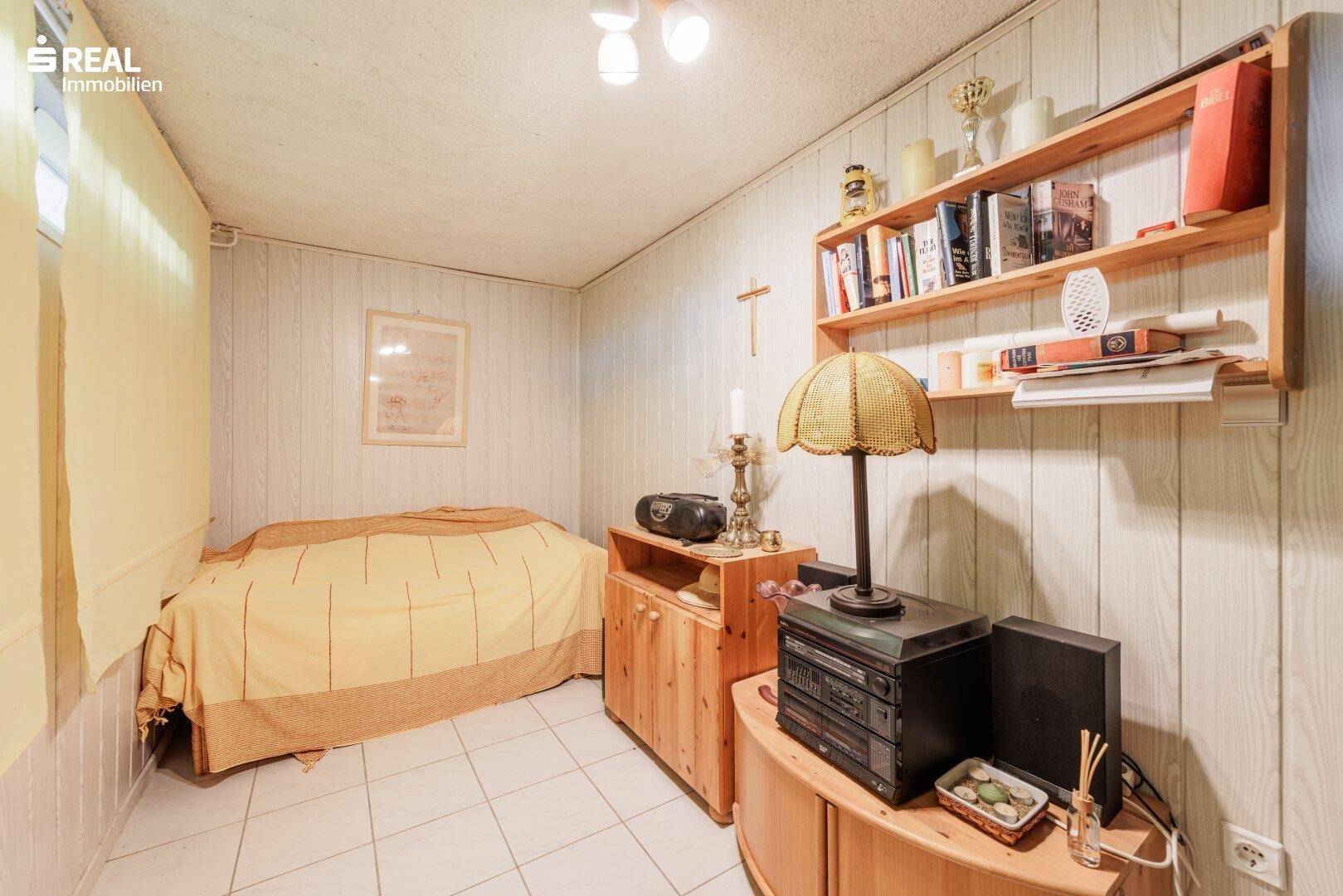 Einfaches Zimmer im Keller mit Bett, Regal und Holzvertäfelung an den Wänden.