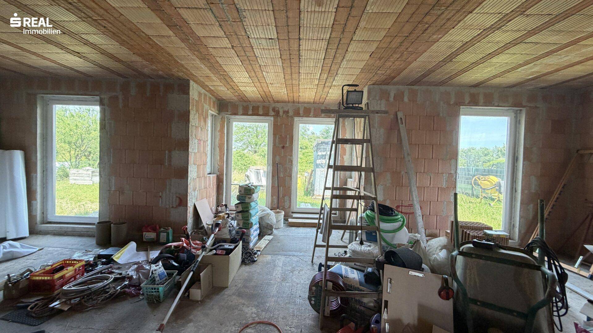 Großer Innenraum im Rohbau mit Ziegelwänden, Holzdecke und Blick ins Grüne.