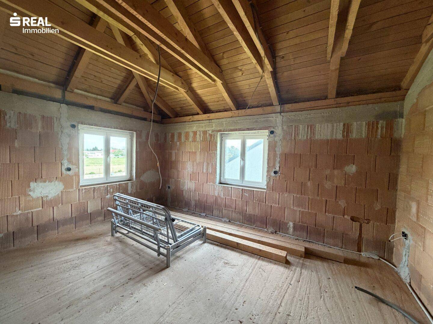 Schlafzimmer im Rohbau mit Holzdachstuhl, Ziegelwänden und zwei Fenstern.
