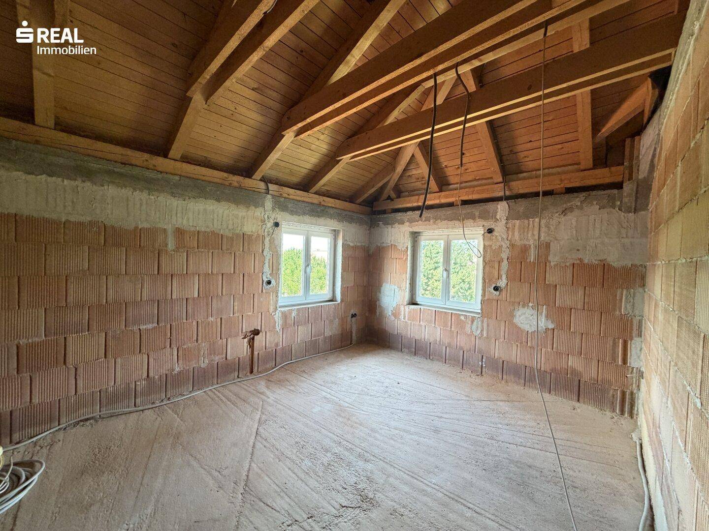 Geräumiges Zimmer im Rohbau mit sichtbarem Dachstuhl und Ziegelwänden.