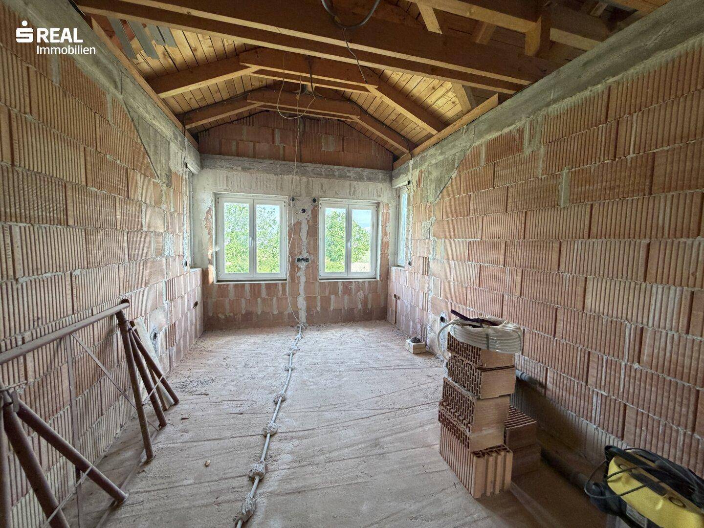 Langer Raum im Rohbau mit Holzdachstuhl, Ziegelwänden und Fenstern.