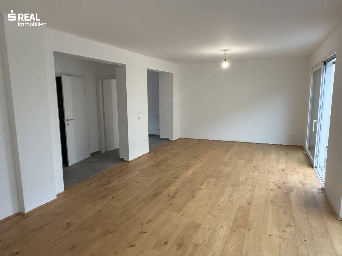 Geräumiges, leeres Wohnzimmer mit hellem Parkettboden und Zugang zu weiteren Räumen.