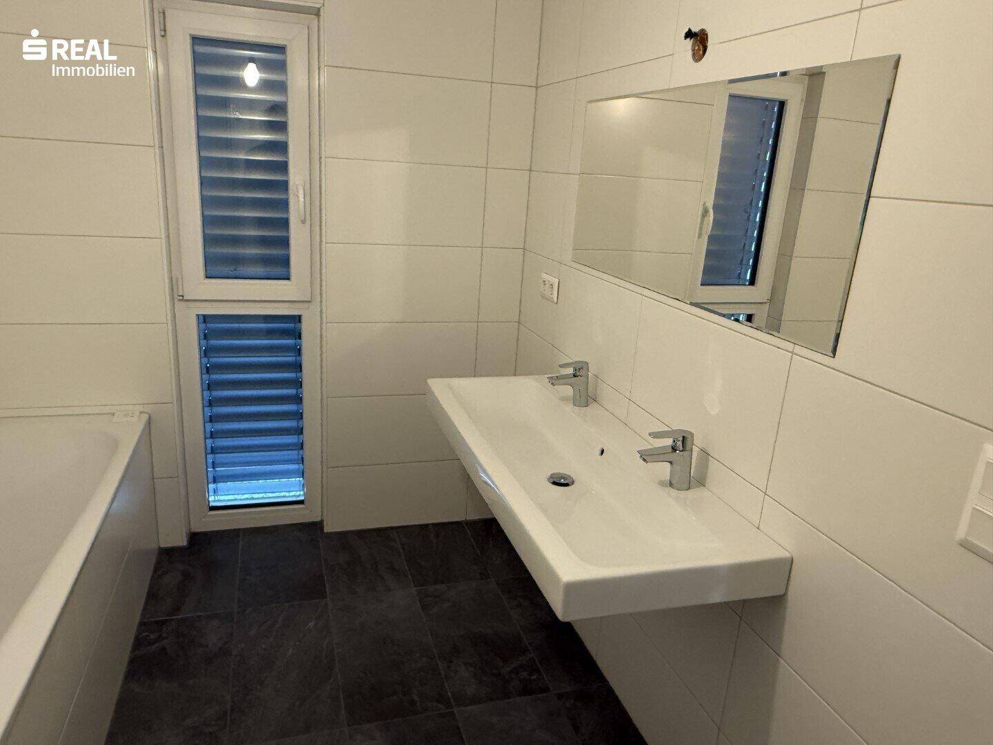 Modernes Badezimmer mit Doppelwaschbecken und Fenster, komplett gefliest.