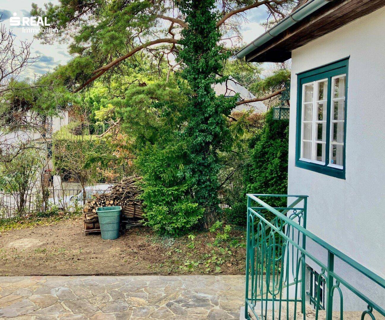 Gepflasterte Terrasse mit grünem Geländer, umgeben von Bäumen und einem Holzstapel.