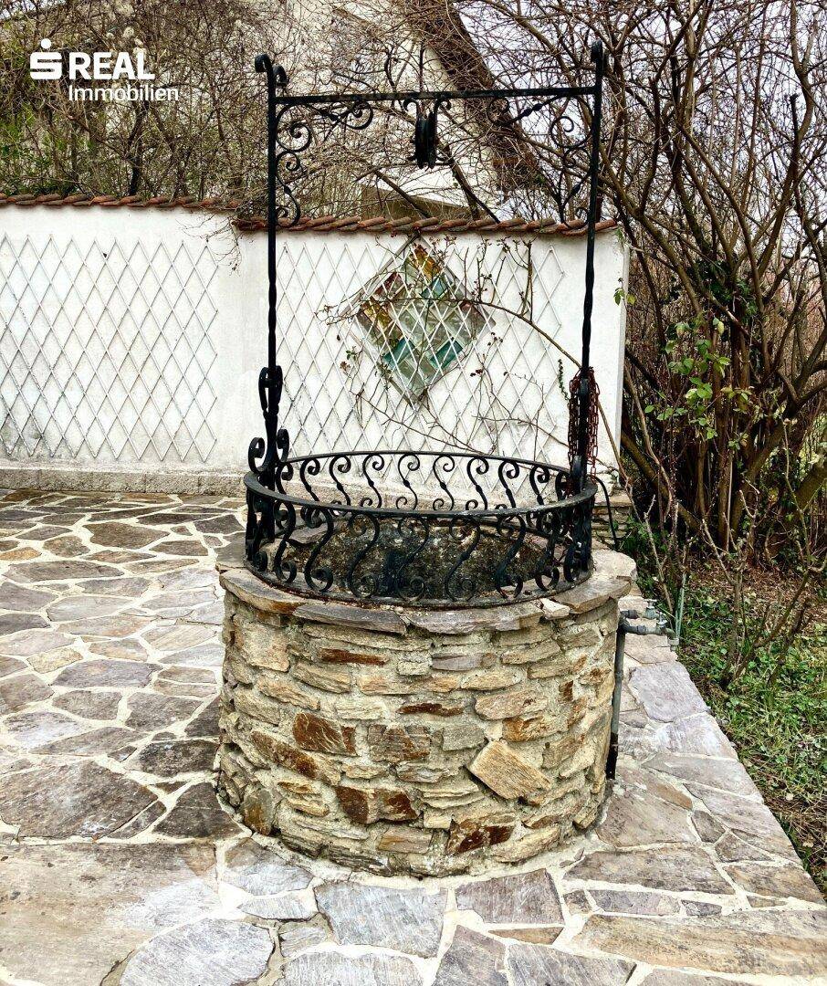 Dekorativer Steinbrunnen mit schmiedeeisernem Aufsatz auf einer gepflasterten Fläche im Garten.