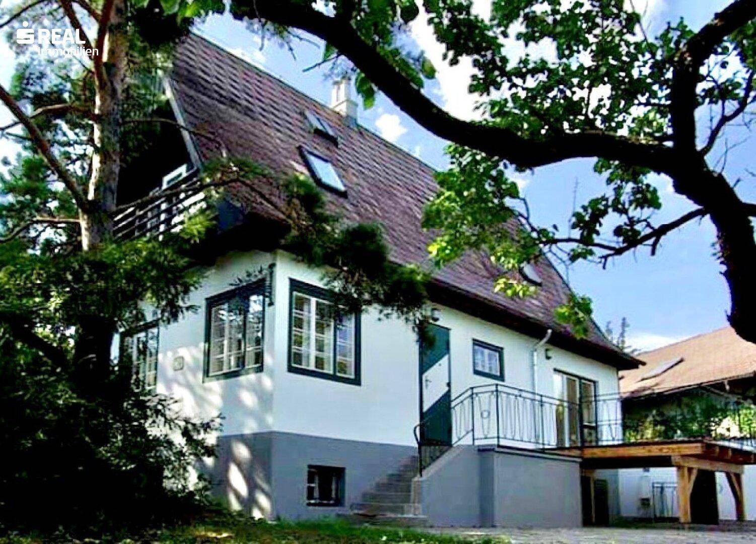 Weißes Haus mit grün umrandeten Fenstern, Satteldach und einer kleinen Terrasse mit Holzunterbau.