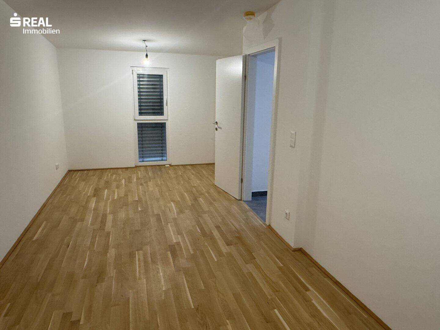Geräumiges Schlafzimmer mit hellem Parkettboden und Fenster mit Jalousien.