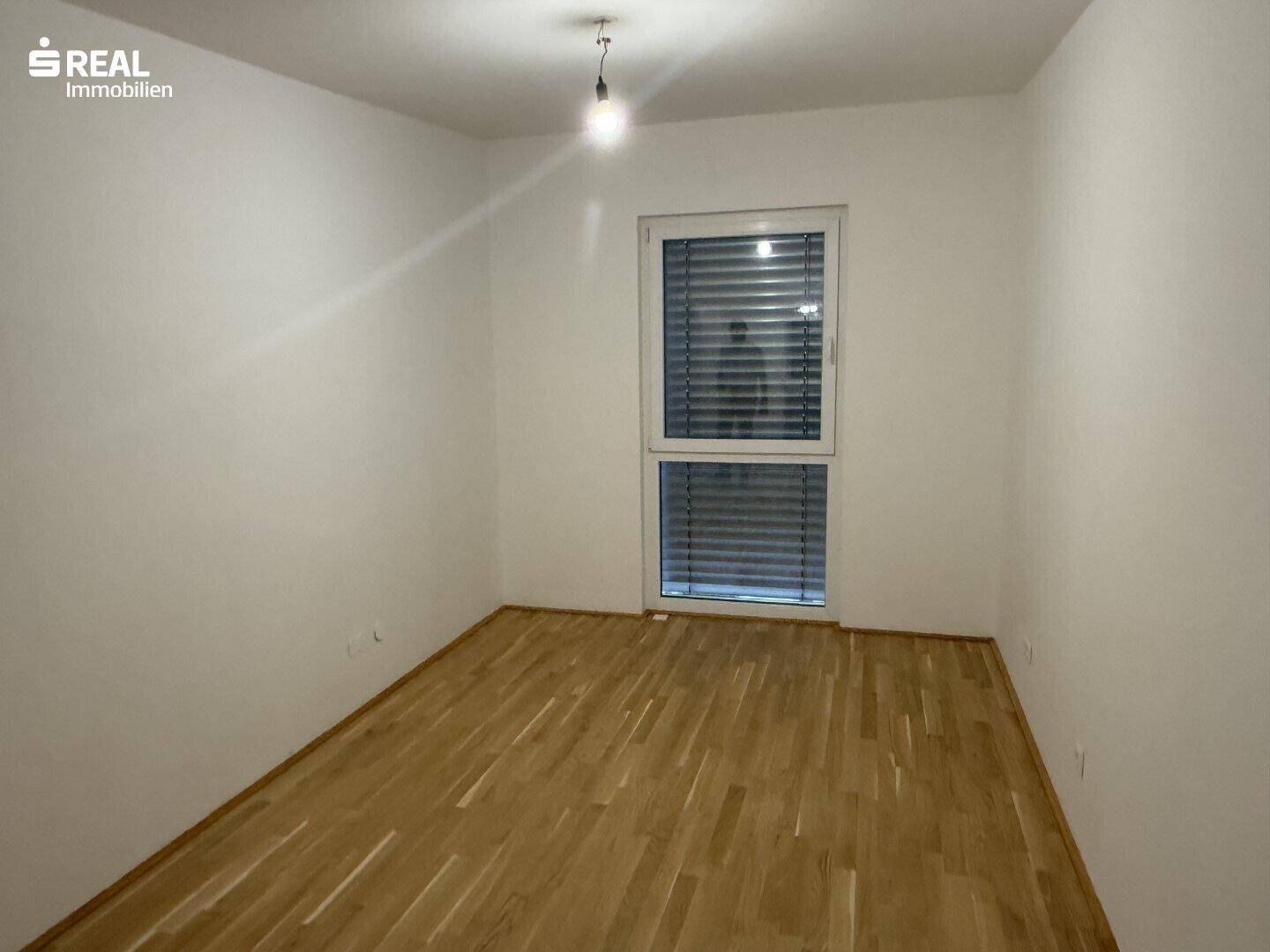 Helles Schlafzimmer mit Parkettboden und großem Fenster mit Außenjalousien.