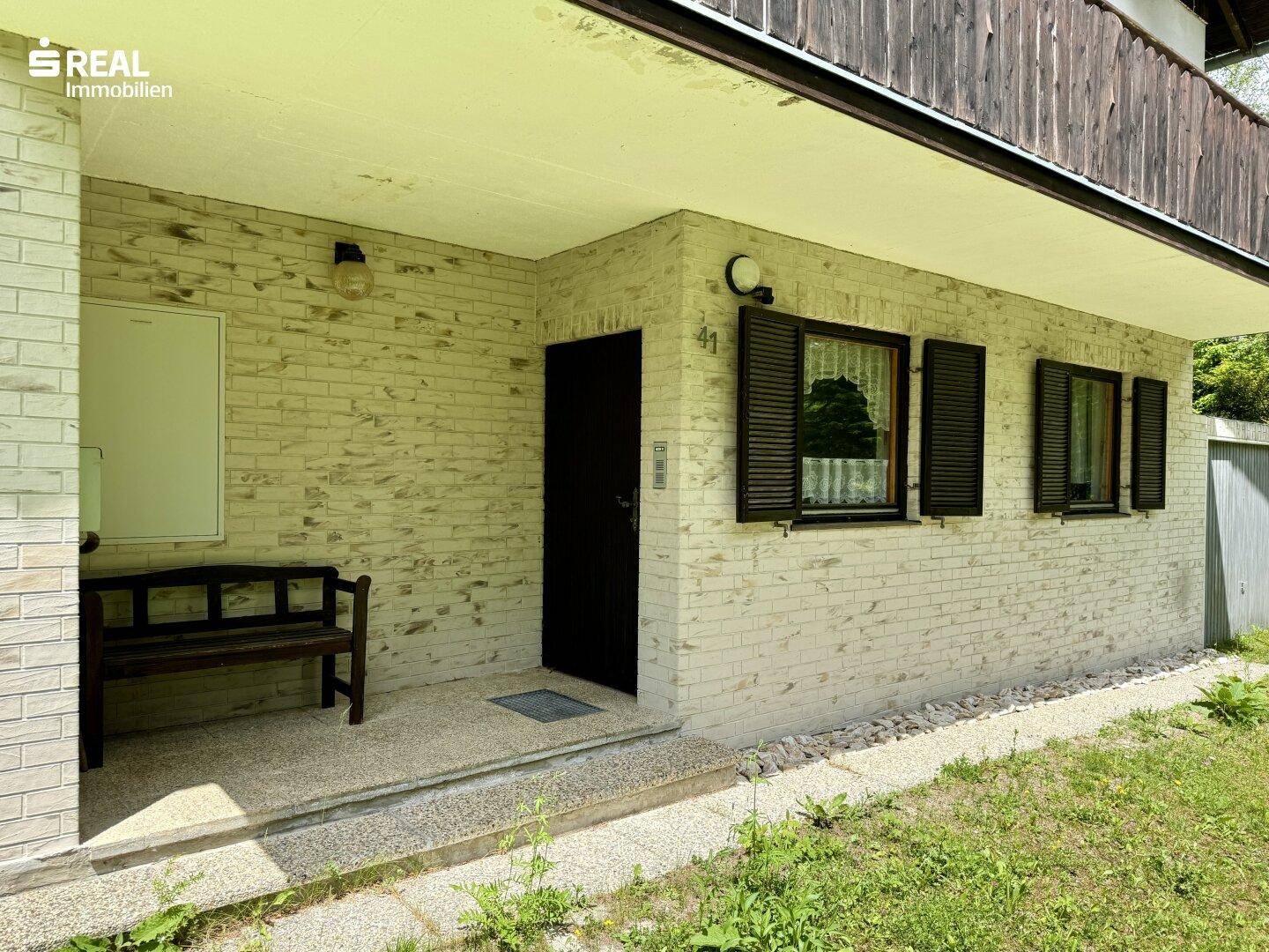 Eingangsbereich des Hauses mit kleiner Bank und Fensterläden, umgeben von Natur.