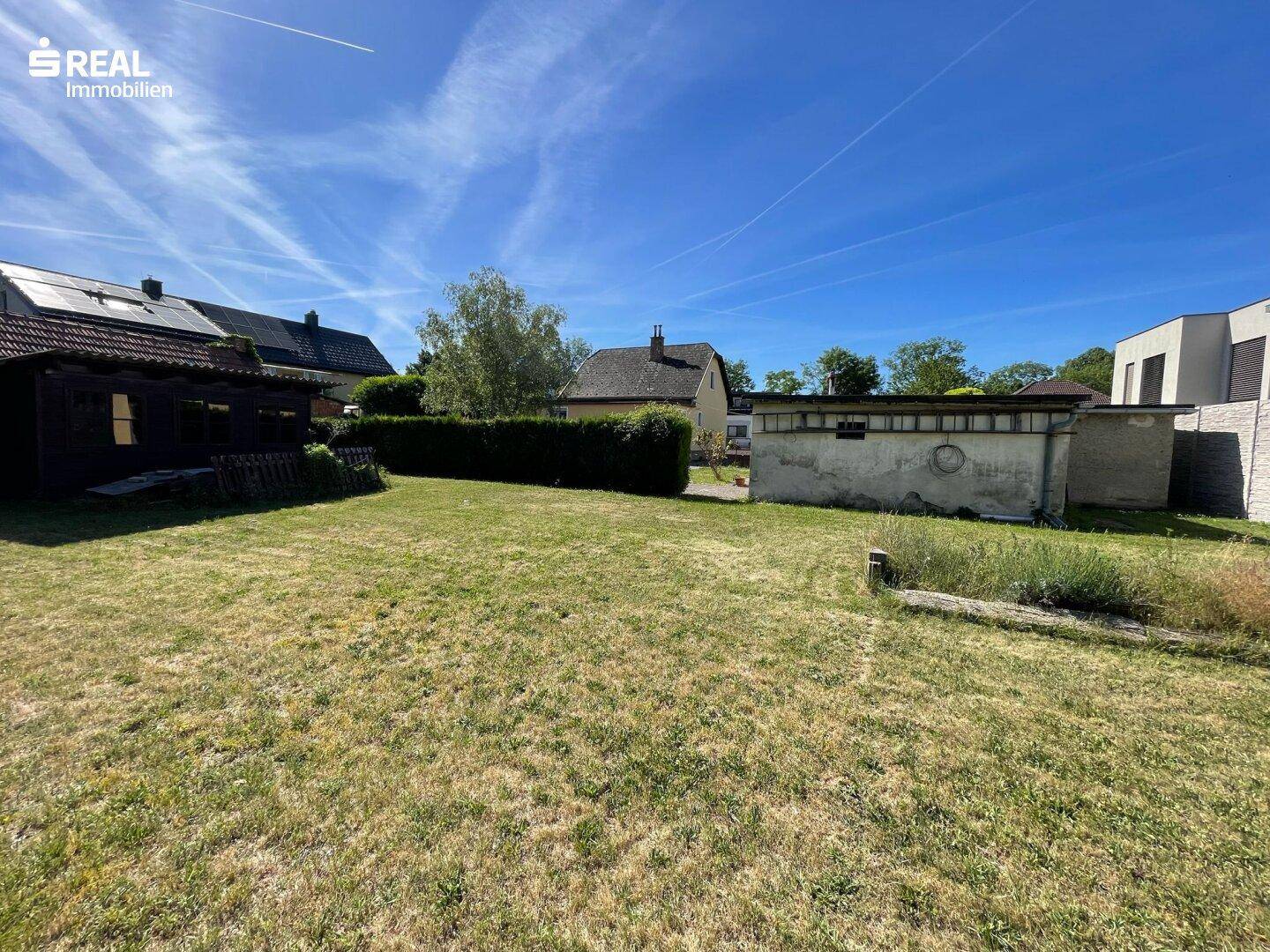 Sonniger Gartenbereich mit Rasen, Hecke und einem kleinen Nebengebäude im Hintergrund.