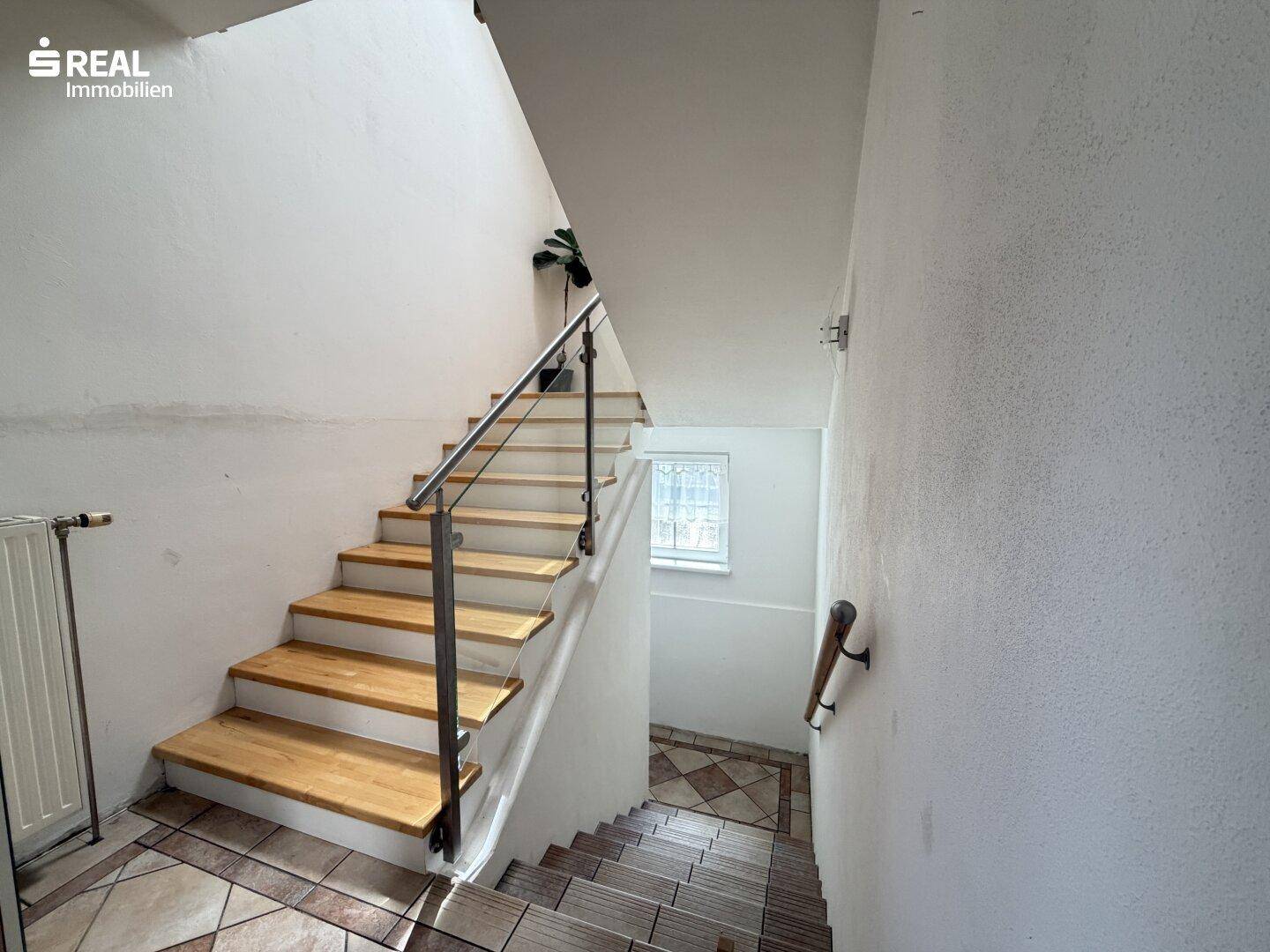 Treppenhaus mit Holzstufen, Glasgeländer und Fliesenboden, gut beleuchtet.