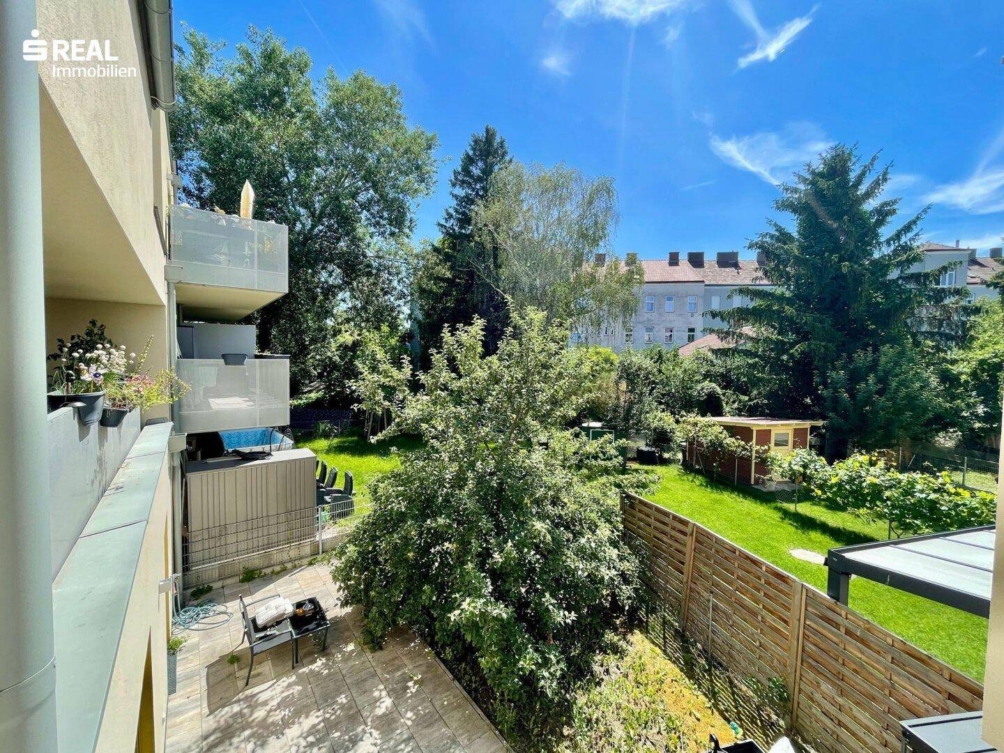 Sonniger Gartenbereich mit Rasenfläche, Bäumen und einem kleinen Gartenhaus, umgeben von einer Holzumzäunung.