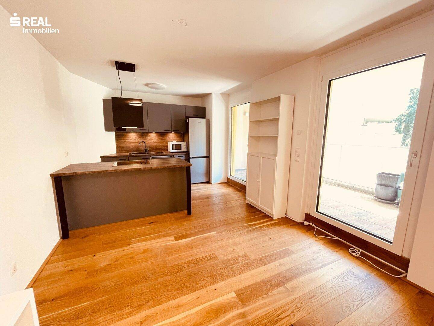 Heller offener Wohnbereich mit moderner Küchenzeile, Holzboden und Zugang zu einem Balkon.