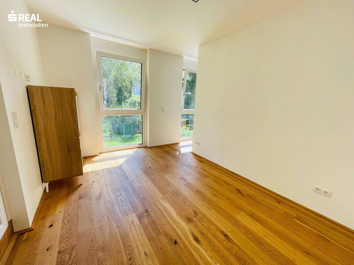 Lichtdurchfluteter Raum mit großen Fenstern, Holzboden und Blick ins Grüne, ideal als Wohn- oder Schlafzimmer.