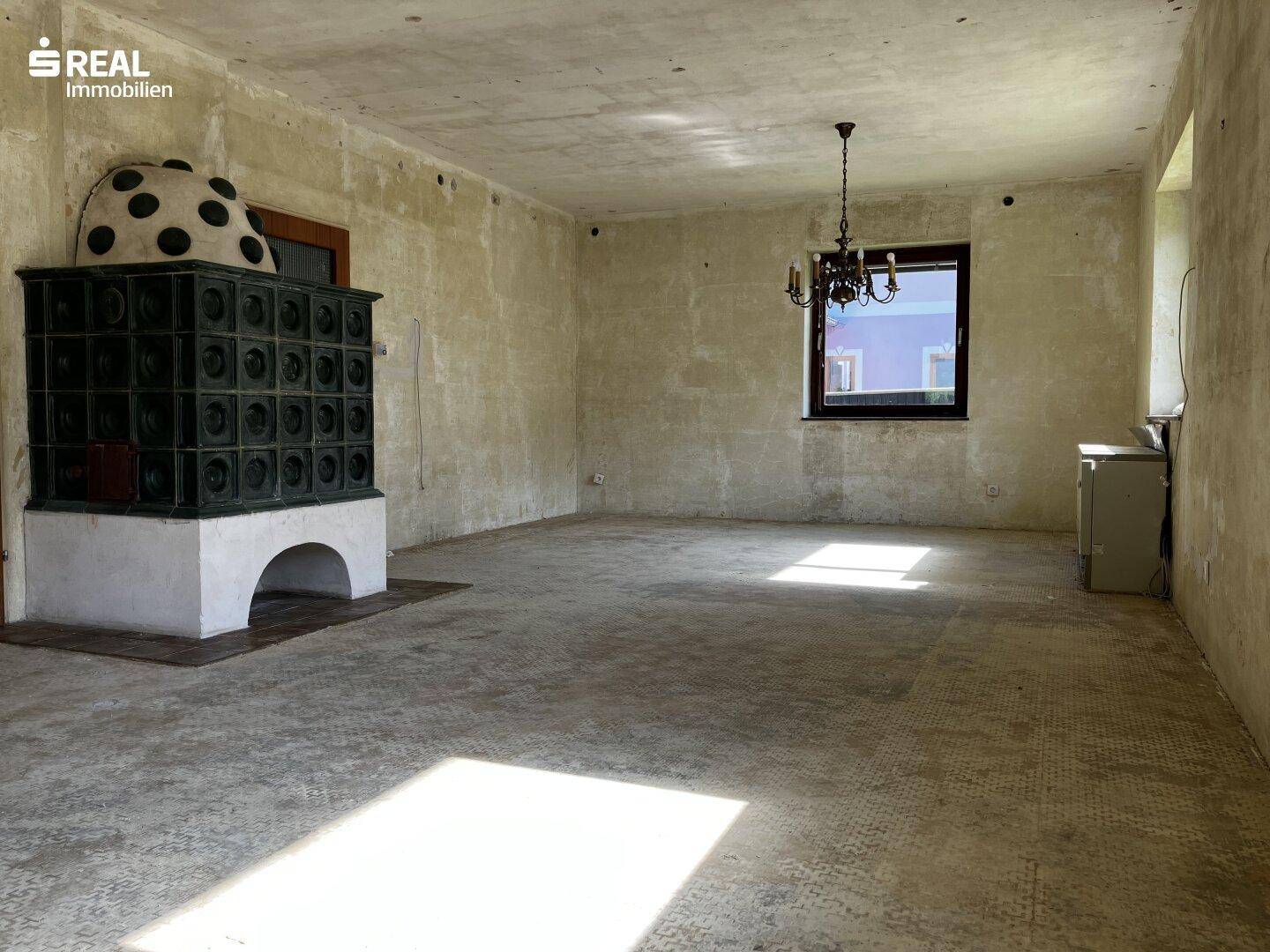 Geräumiges Wohnzimmer mit traditionellem Kachelofen und unfertigem Boden, bereit zur Gestaltung.