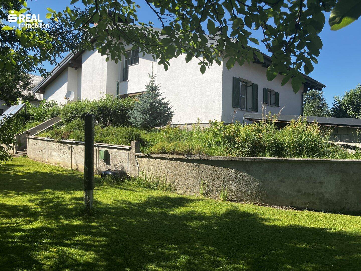 Gartenansicht des Hauses mit terrassiertem Gelände und üppiger Bepflanzung unter blauem Himmel.