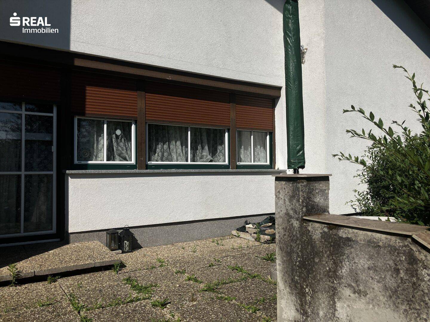 Kleiner gepflasterter Terrassenbereich vor dem Haus mit Fenstern und braunen Rollläden.